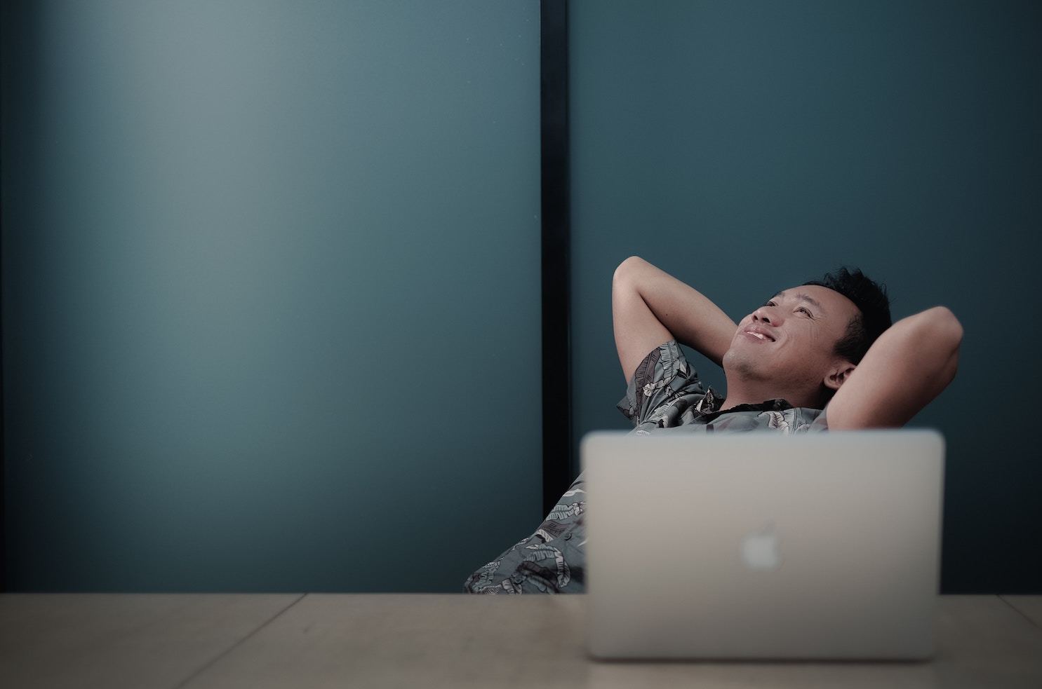 a smiling man tips back in his desk chair, a laptop open in front of him