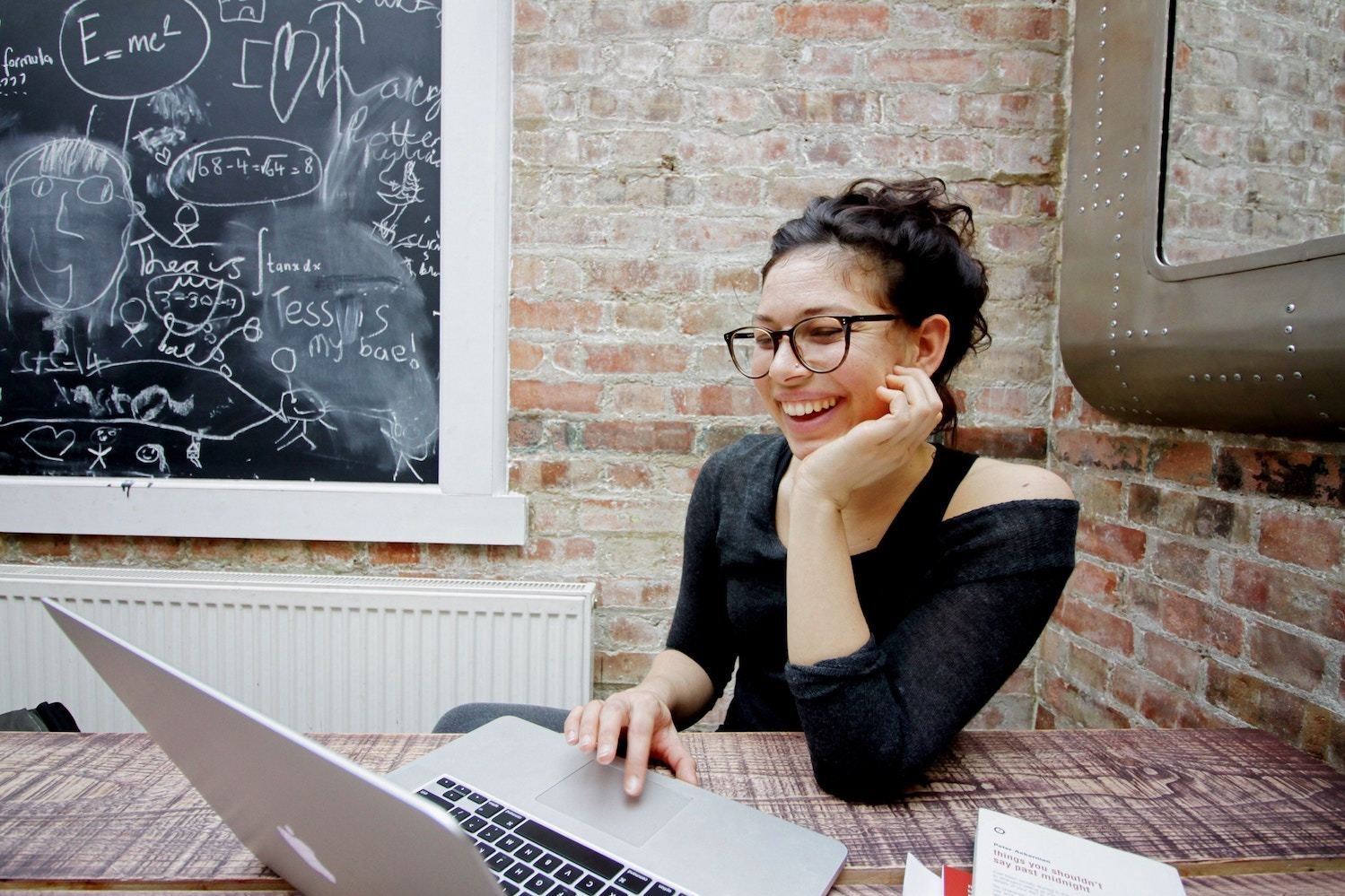 A young woman in glasses laughs while working on her laptop