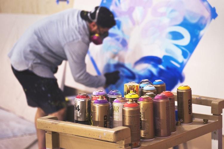 painter paints canvas, with cans of spray paint on a tray in the forefront