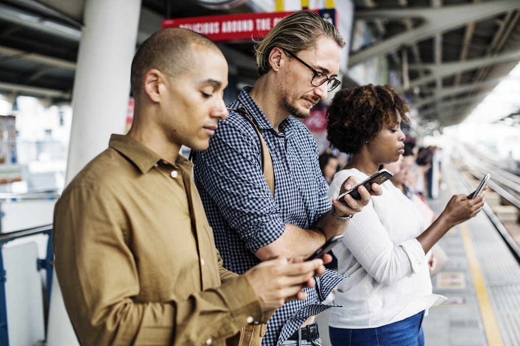 Three people stand side-by-side on a train platform while looking at their phones.