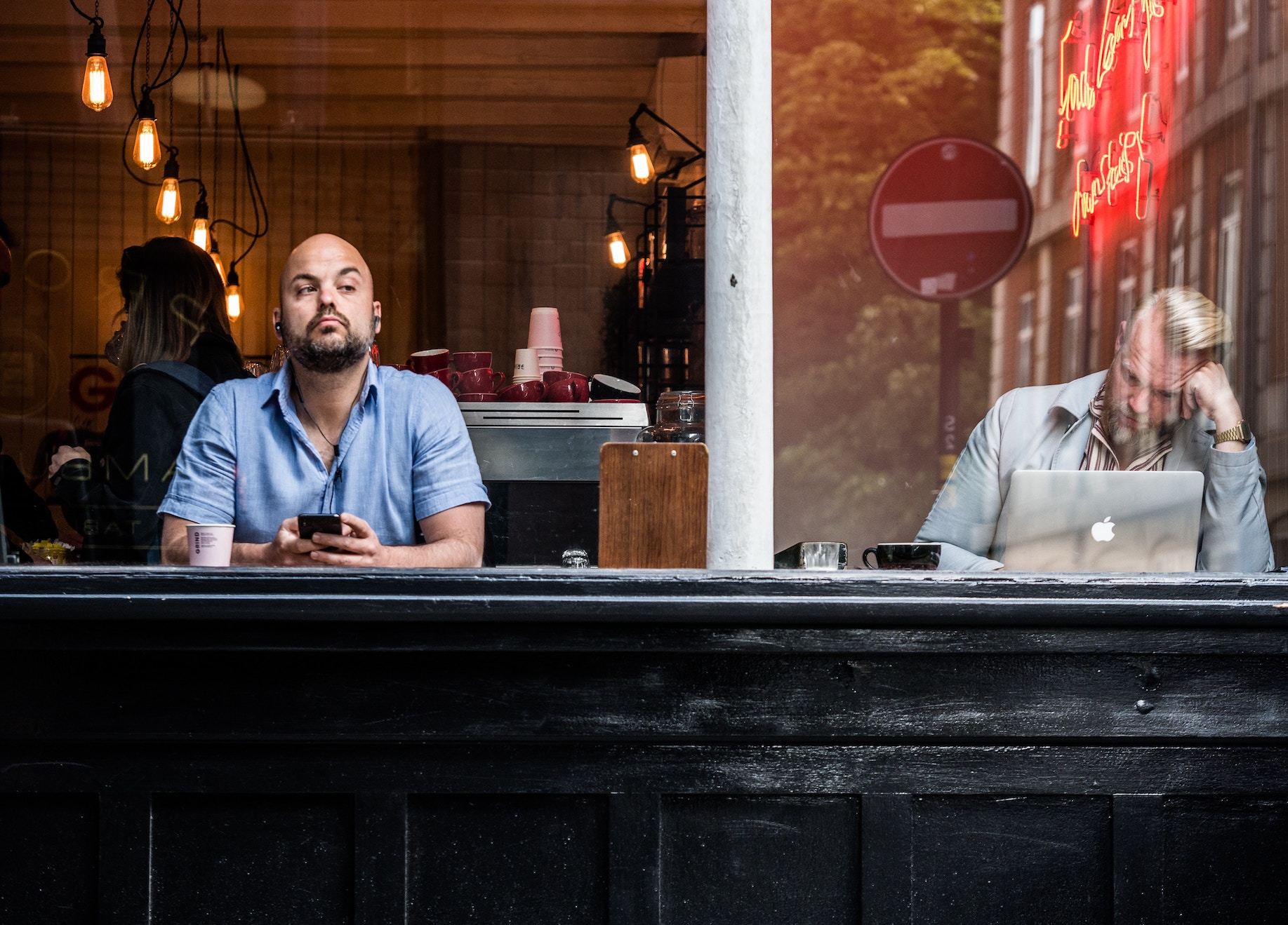 Two men at a cafe counter, one searches on laptop whiel the other looks out to the street