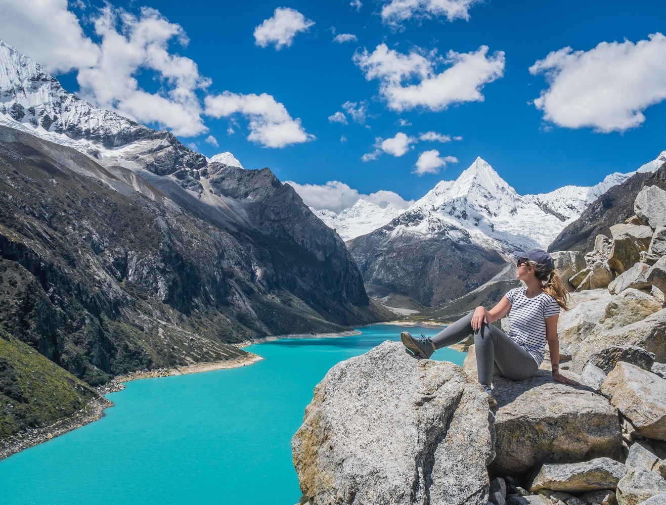 woman sits on rock ledge overlooking green lake surrounded by mountains