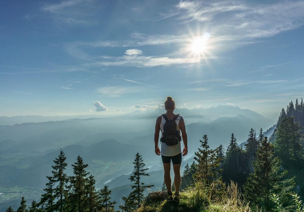 A female hiker pauses at the top of a mountain, surveying the trees and the valley below.