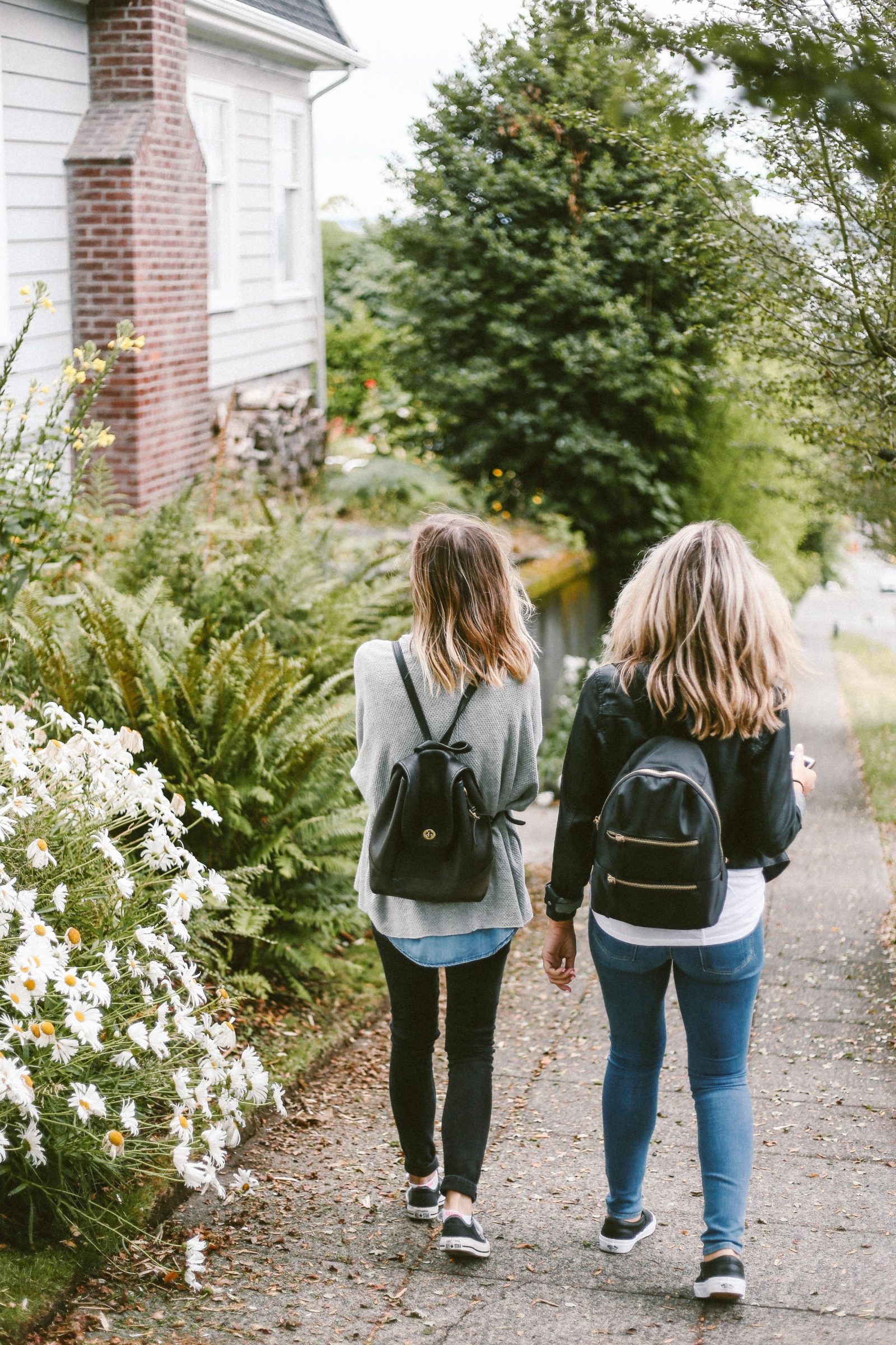 Two young women walk down a sidewalk wearing backpacks.