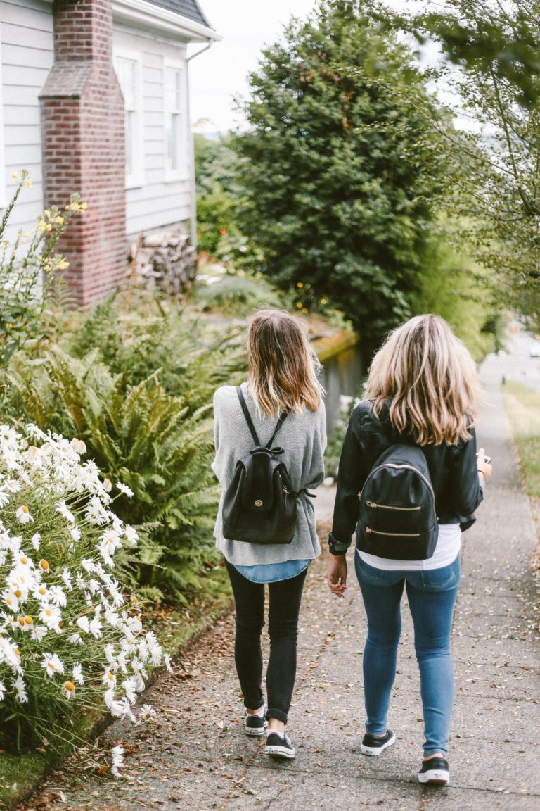 Two young women walk down a sidewalk wearing backpacks.