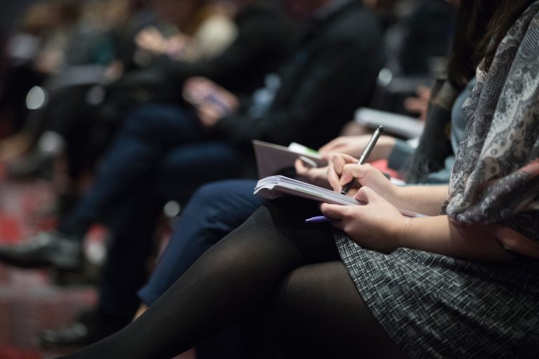 A woman wearing a sweater dress and tights takes copious notes during a live marketing event