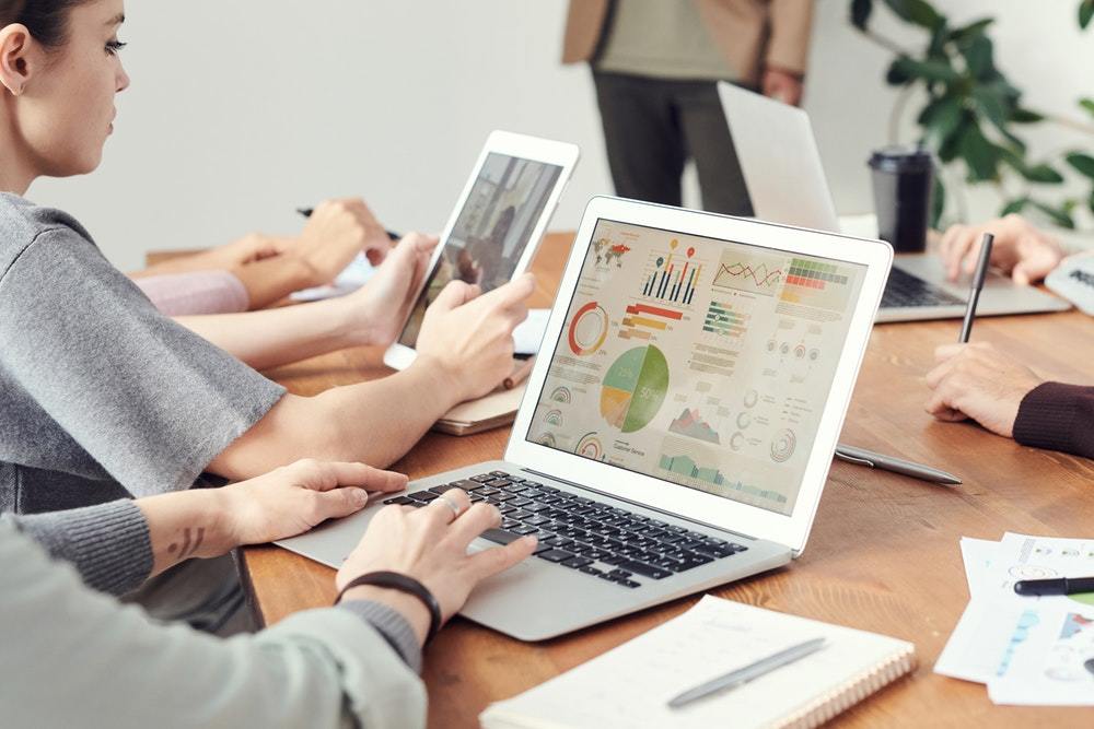 Marketing colleagues are seated around the table in a light, airy room -- one with a laptop displaying analytics, the other with a tablet showing a photo.