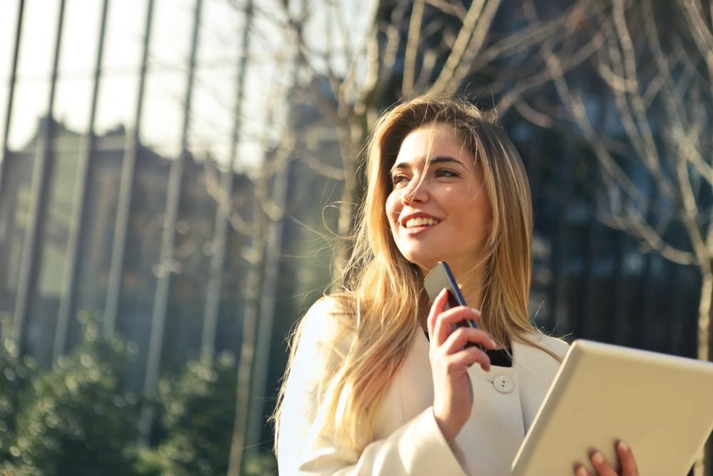 Smiling woman holding up a smartphone and tablet in the afternoon sunlight.