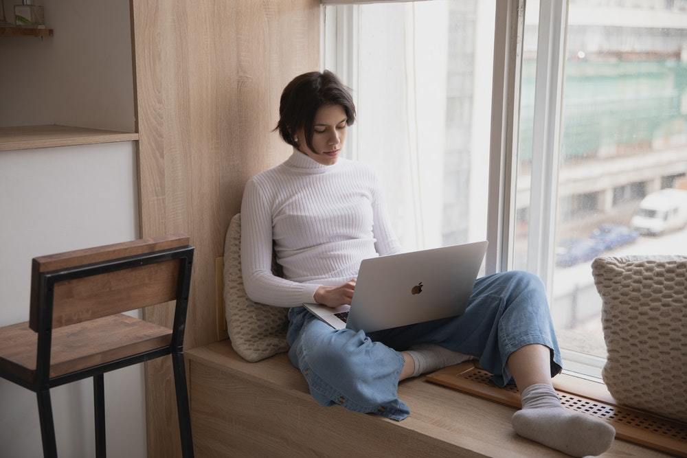 A woman works on a brand resilience plan for her brand on a laptop computer while sitting on a window ledge.