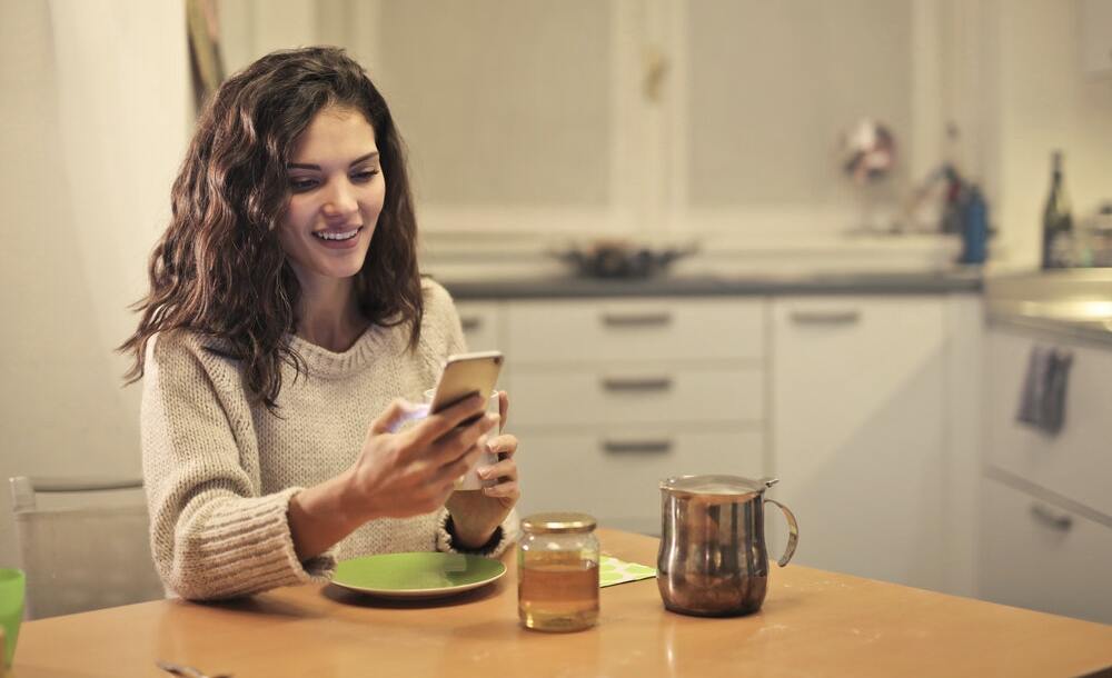A woman wearing a sweater drinks tea and browses content marketing stories on her smartphone.