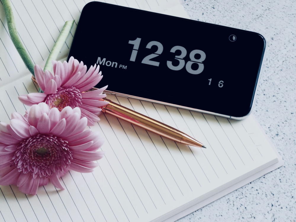 Smartphone with a large clock display placed alongside a brass pen, a blank notepad, and pink Gerbera daisies on a granite desktop.